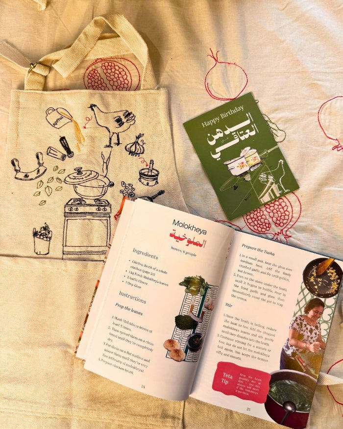 Open Egyptian cookbook and a greeting card beside a cream apron with a hand-drawn molokheya cooking illustration