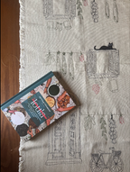 A book titled 'Teta Loula's Cookbook' placed diagonally on a beige linen table runner with black and red illustrated patterns, set on a dark wooden table.