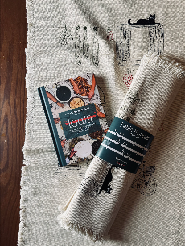 A rolled cream linen table runner with fringed edges and a dark teal paper band reading 'Table Runner'. A hardcover book titled 'Ioula' is placed beside them on a wood surface.