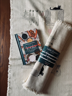 A rolled cream linen table runner with fringed edges and a dark teal paper band reading 'Table Runner'. A hardcover book titled 'Ioula' is placed beside them on a wood surface.